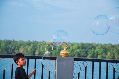 Boy holding wand with bubbles by river against sky