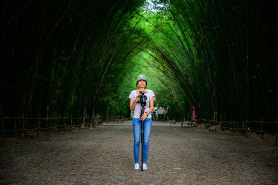 Woman using camera while standing on road against trees