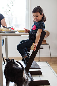 Girl giving bread to dog while sitting at table in house