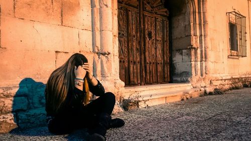 Depressed woman sitting on sidewalk against house in town