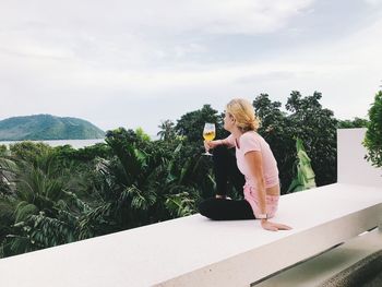 Woman having wine while sitting on wall against sky