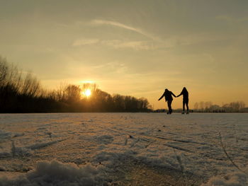 Silhouette of persons skating on lake against sky during sunset