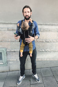 Full length portrait of mid adult man with mobile phone while carrying baby in carrier on sidewalk