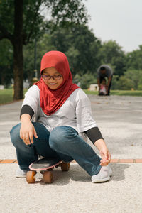 Young girl wearing hijab is running while holding pennyboard in the park. view from behind.