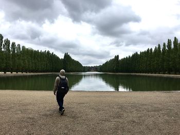 Rear view of man by pond amidst trees at parc de sceaux against cloudy sky