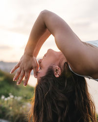 Side view of calm young female with long brown hair doing backbend asana with closed eyes and prayer hands near face while practicing yoga at seaside at sunset