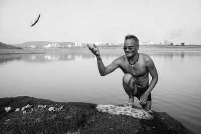 Shirtless fisherman throwing fish from net while kneeling on lakeshore