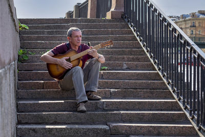 Adult man plays six-string classical acoustic guitar outdoors while sitting on a granite staircase 