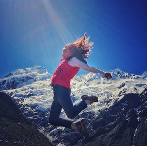 Low angle view of young woman jumping over snowcapped mountain against blue sky