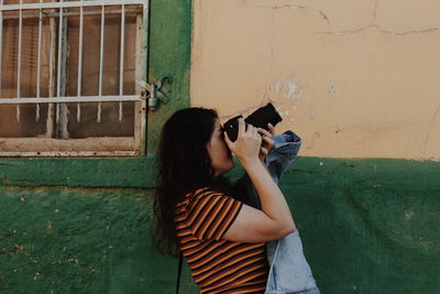 Woman photographing while standing by wall outdoors