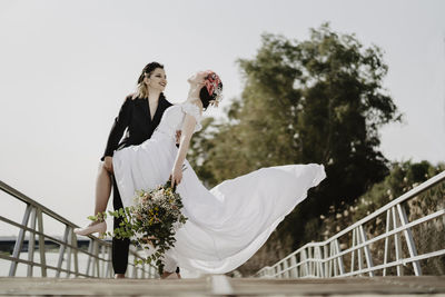 Ground level of cheerful woman in black suit smiling and supporting fiancee in white dress tilting back while standing on bridge on wedding day in park