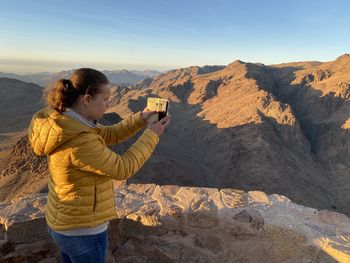 Side view of man photographing with camera while standing on mountain against sky