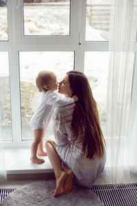 Mother in a white robe sits with a child a blonde daughter at a large window of the house person