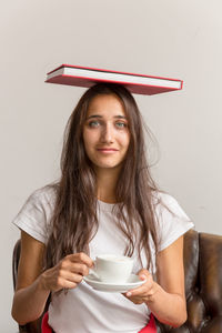 Portrait of woman holding coffee cup with book on head