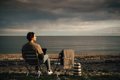 Thoughtful man using laptop while sitting at beach against cloudy sky