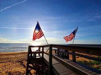 Two american flags resting on a wooden walkway over the beach with the ocean behind them