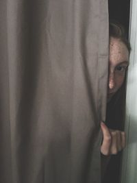 Close-up portrait of teenage girl peeking through curtain at home