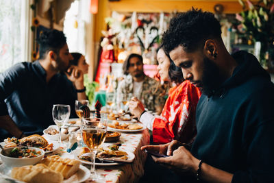 Young man text messaging through smart phone while sitting with friends during brunch party at restaurant