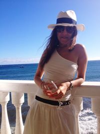 Woman in sunglasses and hat holding wineglass against sea and sky on sunny day