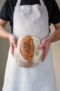 Midsection, bakers hands holding a round shaped loaf of organic sourdough bread in front of him.