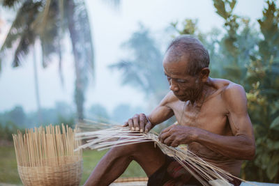 Shirtless man weaving wicker basket against trees