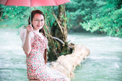 Portrait of smiling young woman holding umbrella sitting by river