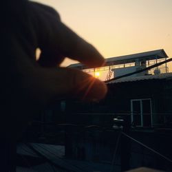 Person hand by building against sky during sunset