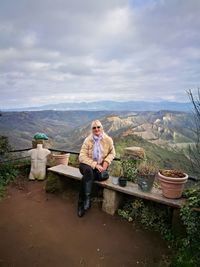 Full length portrait of woman sitting on bench against landscape and sky