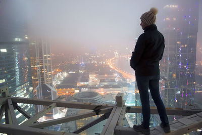 Rear view of woman standing by railing against illuminated buildings in city