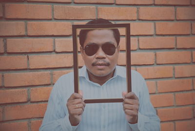 Portrait of man holding picture frame while standing against brick wall