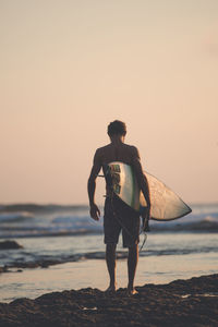 Rear view of man with surfboard standing at beach against clear sky during sunset