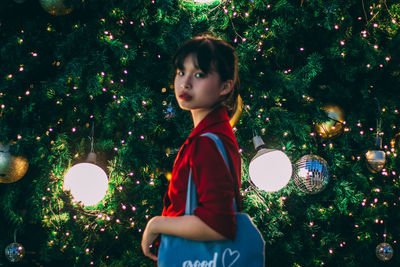 Portrait of woman standing against illuminated christmas tree at night