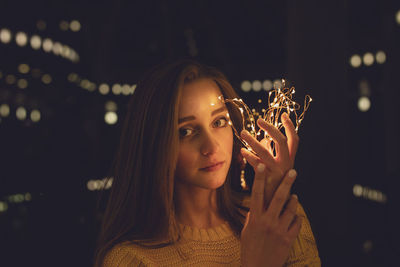 Portrait of young woman holding illuminated string lights in darkroom