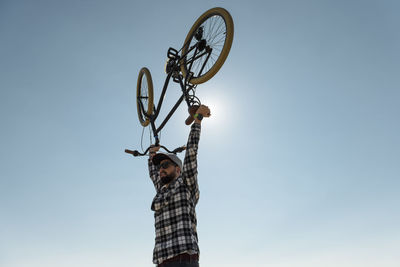 Low angle view of man with arms raised carrying bicycle while standing against clear blue sky during sunny day