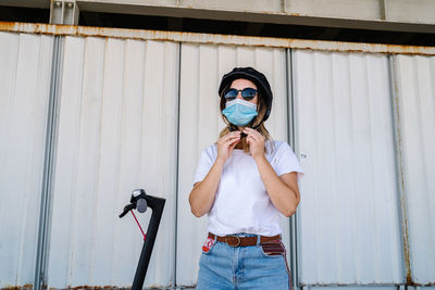 Young female rider in medical mask and sunglasses putting on protective helmet while preparing for riding electric scooter