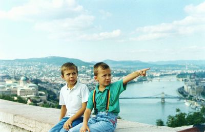 Boy sitting with brother pointing on bridge over river amidst city