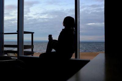 Silhouette woman sitting by sea against sky seen through window