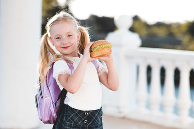 Cute child girl 5-6 year old holding hamburger outdoors. wearing school bag and uniform close up