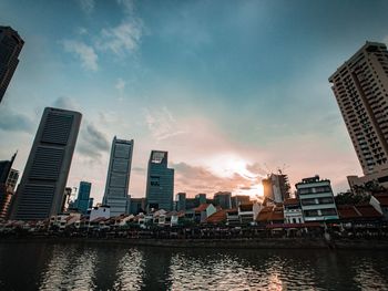 Modern buildings by river against sky during sunset