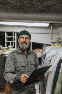 Portrait of bearded tailor with clipboard standing by clothing rack in workshop