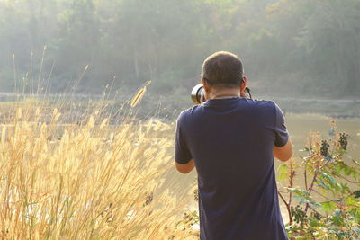 Rear view of man photographing while standing amidst plants by lake