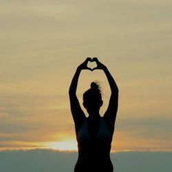 Silhouette woman making heart shape with arms raised against sky during sunset