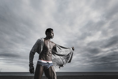 Low angle view of muscular man standing in underwear at beach against cloudy sky