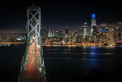 Illuminated bridge over river against buildings at night