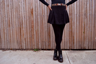 Low section of woman standing on sidewalk with legs crossed at ankle against wooden fence