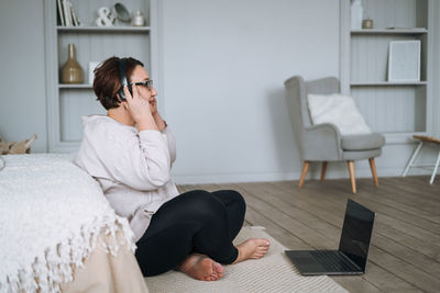 Middle aged plus size smiling brunette woman in home wear listening music in headphones using laptop 