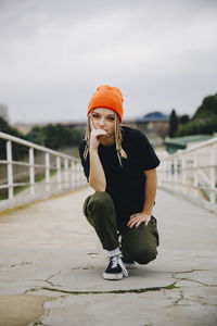 Confident young woman wearing orange knit hat posing on footbridge against sky