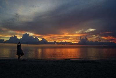 Silhouette woman doing tree pose while standing at beach against sky during sunset