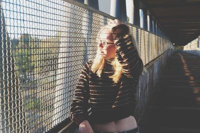 Young woman looking through chainlink fence while standing on bridge
