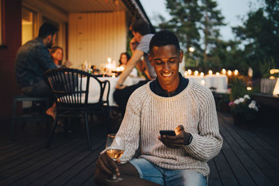 Smiling young man using mobile phone while friends in background during dinner party